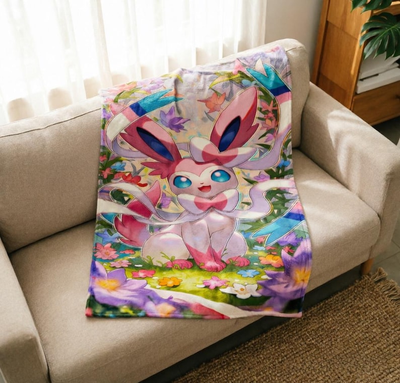 May include: A soft throw blanket featuring a colorful illustration of the pink and white Sylveon character from the Pok&eacute;mon franchise, surrounded by a vibrant garden of purple, yellow, and white flowers with green foliage and blue ribbons.