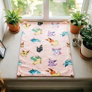 May include: A rectangular light pink fleece blanket featuring a repeating pattern of various colorful cartoon creatures, including yellow, blue, purple, and orange characters with distinct ears and tails, scattered across the soft fabric surface.