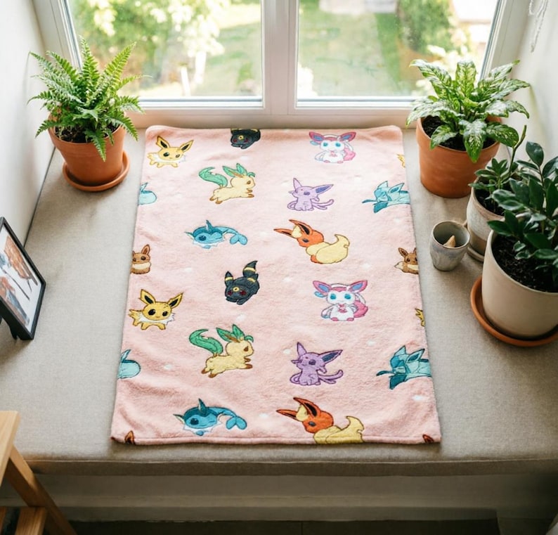 May include: A rectangular light pink fleece blanket featuring a repeating pattern of various colorful cartoon creatures, including yellow, blue, purple, and orange characters with distinct ears and tails, scattered across the soft fabric surface.