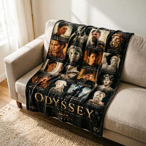 May include: A black fleece throw blanket featuring images of characters from the Odyssey. The blanket has a gold title that reads "The Odyssey" with the text "Not all who wander are lost" and "IMAX 2026". The blanket is draped over a beige sofa.