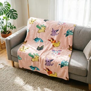 May include: A soft, light pink throw blanket featuring a repeating pattern of various colourful Eevee evolution characters. The blanket displays multiple small, illustrated creatures in shades of yellow, blue, purple, and orange scattered across the fabric.