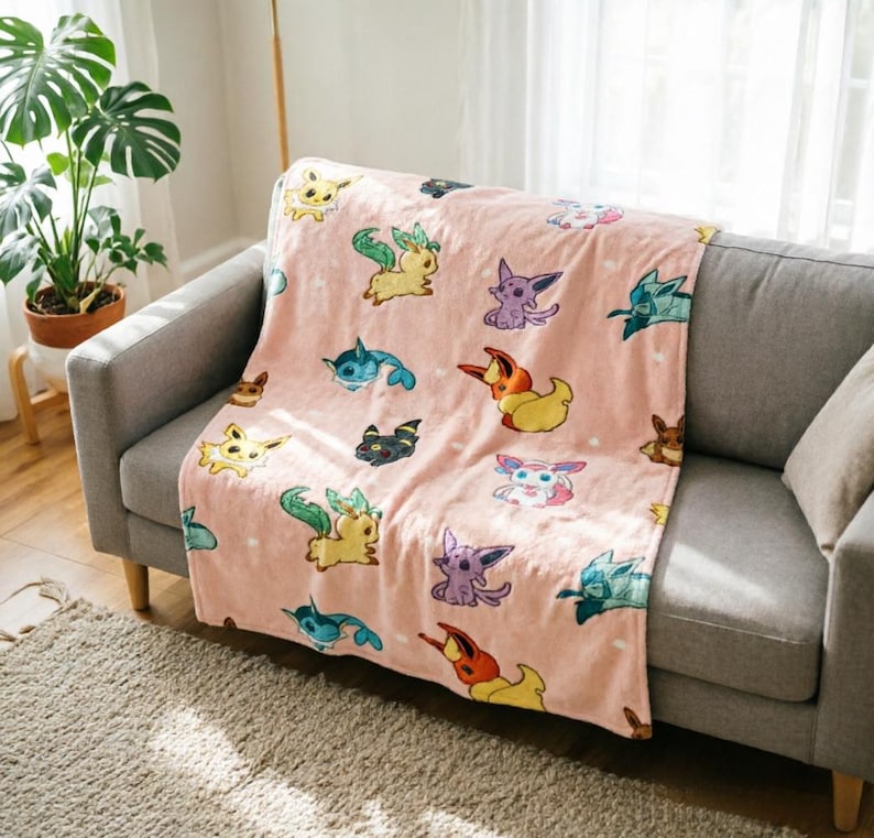 May include: A soft, light pink throw blanket featuring a repeating pattern of various colorful Eevee evolution characters. The blanket displays multiple small, illustrated creatures in shades of yellow, blue, purple, and orange scattered across the fabric.