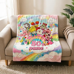 Op de afbeelding: Een kleurrijke deken met een ontwerp van tekenfilmfiguren, een regenboog en wolken. De deken heeft de tekst "made in the 80s" en een ruimte voor een naam. De deken ligt over een bank.