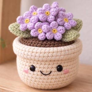 May include: A crocheted flower pot with a smiling face. The pot is cream-colored and contains a brown crocheted base with purple flowers and green leaves. The flowers have yellow centers and the pot has black eyes and pink cheeks.