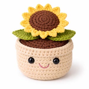 Pode incluir: Um girassol de crochê num vaso sorridente. O girassol tem pétalas amarelas e um centro castanho. O vaso é bege com olhos pretos e um sorriso rosa. As folhas são verdes.