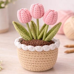 Può includere: Un vaso di fiori all'uncinetto con tre tulipani rosa, steli e foglie verdi. Il vaso beige ha un bordo smerlato bianco. I tulipani sono in terra all'uncinetto marrone. Decorazione floreale fatta a mano.