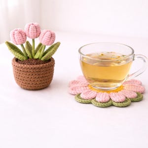 Könnte beinhalten: Ein gehäkelter Blumentopf mit drei rosa Tulpen und grünen Blättern steht neben einer durchsichtigen Teetasse auf einem gehäkelten Blumenuntersetzer in Rosa, Gelb und Grün. Der Tee hat eine hellbernsteinfarbene Farbe.