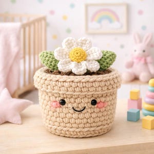 Puede incluir: Una maceta de ganchillo con una cara sonriente, mejillas rosadas y ojos negros. La maceta es beige y contiene una margarita blanca con un centro amarillo y hojas verdes. La maceta está sobre una superficie de madera.