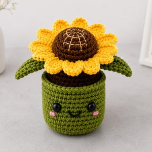 Pode incluir: Um girassol de crochê em um vaso verde com um rosto sorridente. O girassol tem pétalas amarelas, um centro marrom e folhas verdes. O vaso é verde com olhos pretos e bochechas rosadas.