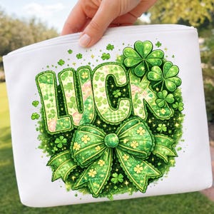 Puede incluir: Bolsa blanca con cremallera con un diseño "LUCK" verde y amarillo. El diseño incluye un lazo verde, tréboles y la palabra "LUCK" en una fuente verde texturizada. La bolsa es sostenida por una persona.