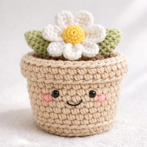 Puede incluir: Una maceta de ganchillo con una cara sonriente, mejillas sonrojadas y una margarita blanca con centro amarillo. La maceta es beige y la flor tiene hojas verdes. El artículo hecho a mano es una pieza decorativa encantadora.