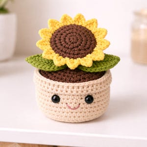 May include: A crocheted sunflower in a pot. The sunflower has yellow petals and a brown center. The pot is beige with a smiling face, black eyes, and a pink mouth. Green leaves are visible beneath the flower.