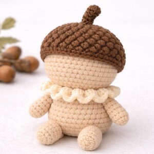 May include: A crocheted acorn doll with a brown cap and a cream-colored body, arms, and legs. The doll has a ruffled collar and is sitting. The acorn cap is textured, and the doll is made of soft yarn.