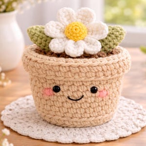 Pode incluir: Um vaso de flores de crochê com um rosto sorridente, bochechas rosadas e olhos pretos. O vaso bege contém uma margarida branca com centro amarelo e folhas verdes. O vaso está sobre uma toalha de renda branca.