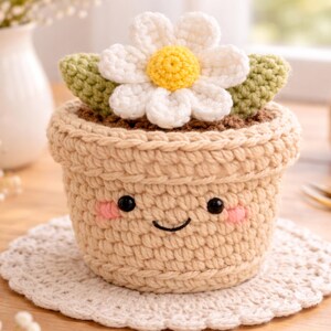 May include: A crocheted flower pot with a smiling face, blush cheeks, and a white daisy with a yellow center. The pot is beige, and the flower has green leaves. The pot sits on a white, crocheted doily.