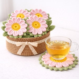 May include: A crocheted flower basket with four pink and yellow daisy-shaped toppers. A matching coaster holds a glass teacup. The basket is brown with a cream-colored lace ribbon and bow.