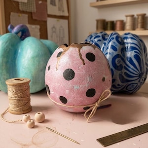 May include: A collection of decorative pumpkins in various colors and patterns. One pumpkin is light blue, another is pink with black polka dots, and a third is blue with white swirls. Crafting supplies, including twine, wooden beads, and a needle, are also present.