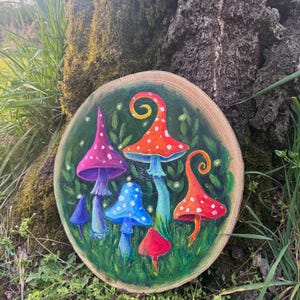 May include: A round wooden plaque featuring a whimsical painting of colorful mushrooms. The mushrooms are painted in shades of red, orange, blue, and purple, with white polka dots. The background is green, with grass and leaves.
