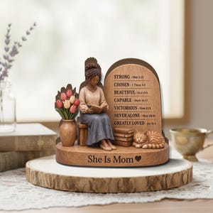May include: A wooden figurine of a woman reading, with a vase of tulips and a sleeping cat. The figurine has the words "She Is Mom" and inspirational phrases like "Strong" and "Chosen" engraved on it. The figurine is on a wooden base.