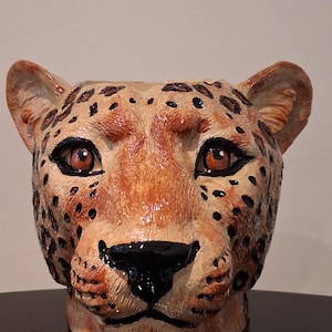 Puede incluir: Escultura de cerámica con forma de cabeza de leopardo con detalles. La escultura tiene un color base beige con manchas negras y ojos marrones. La nariz es negra y las orejas son de color beige. Es un objeto decorativo.