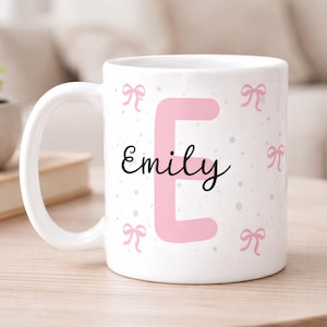 Può includere: Tazza in ceramica bianca con la lettera "E" rosa e il nome "Emily" in corsivo nero. La tazza è decorata con fiocchi rosa e piccoli puntini grigi. La tazza è su una superficie di legno chiaro.