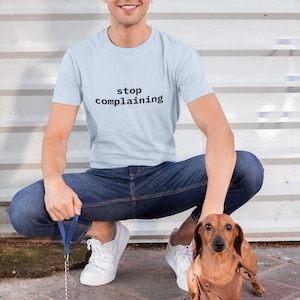 May include: A light blue t-shirt with the words "stop complaining" in black. The shirt is paired with blue jeans and white trainers. A brown dachshund dog wearing a leather harness is also present.