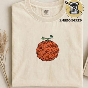 Op de afbeelding: Crèmekleurig T-shirt met een geborduurd oranje fruitontwerp en groene stengel. Het shirt heeft een ronde hals en korte mouwen. Het woord "EMBROIDERED" is op het shirt gedrukt.