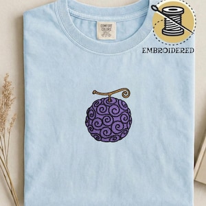 May include: Light blue t-shirt with an embroidered purple fruit design. The fruit has a brown stem and a swirling pattern. The shirt also has the word "EMBROIDERED" and a logo of a needle and thread.