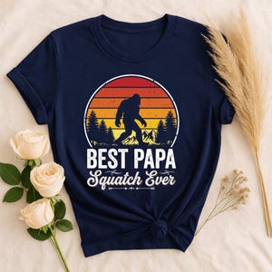 Può includere: Una t-shirt blu navy a maniche corte con una grafica che mostra la sagoma di Bigfoot che cammina davanti a un tramonto con pini e montagne. Il testo recita BEST PAPA Squatch Ever in caratteri bianchi, in grassetto e corsivi.
