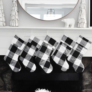 May include: A fireplace mantel decorated for the holidays. Six black and white buffalo check stockings hang above the fireplace. A round mirror and decorative items complete the scene. A sign reads, "In a world where you be anything, be kind."