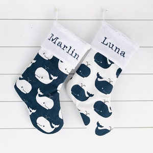 Könnte beinhalten: Zwei Weihnachtsstrümpfe mit weißen Walmustern auf marineblauem und weißem Hintergrund. Die Strümpfe haben weiße Manschetten mit den Namen "Marlin" und "Luna" in Marineblau gestickt. Sie hängen an einer weißen Wand.