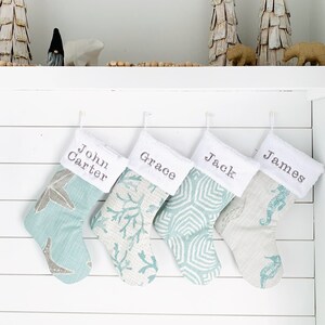 Puede incluir: Cuatro calcetines navideños personalizados con nombres bordados en puños blancos. Los calcetines presentan varios diseños de temática marina en tonos azules y grises. Árboles de madera decorativos y figuras en la repisa superior.