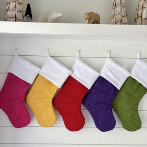 Puede incluir: Seis calcetines navideños coloridos en tonos rosa, amarillo, rojo, morado y verde, cada uno con un puño blanco. Cuelgan de ganchos transparentes en una pared blanca con listones horizontales. Objetos decorativos en un estante.