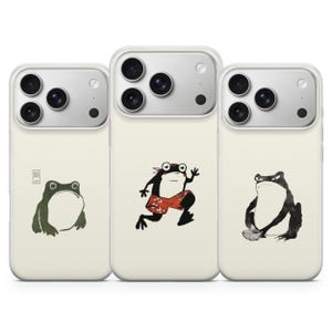 May include: Three phone cases with quirky frog illustrations. The cases have a light cream background and a clear camera lens protector. The frog designs include a green frog, a frog in red shorts, and a grumpy frog.