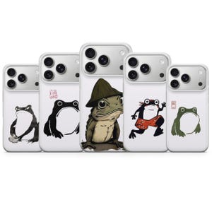 May include: A collection of white phone cases featuring various frog illustrations. The designs include a black and white frog, a frog wearing a hat, and a frog in red shorts. The cases have a glossy finish and a camera cutout.