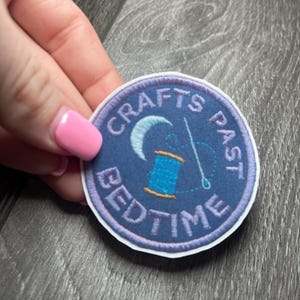 May include: A round, navy blue patch with white trim featuring the words "Crafts Past Bedtime." The patch includes a crescent moon, a spool of thread, and a needle. The patch is held by a hand with pink nail polish.