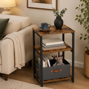 May include: A three-tier side table with a black metal frame and rustic brown wood-grain shelves. The top shelf holds a blue mug on a saucer and a black vase. The middle shelf contains stacked books, and the bottom shelf holds a gray fabric bin.