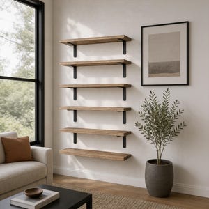 May include: A set of six wall-mounted shelves made of light brown wood with visible grain, each supported by two black metal brackets. The shelves are arranged vertically in a column on a plain wall, positioned next to a framed rectangular art piece.