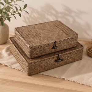 May include: Two rectangular storage boxes crafted from woven brown seagrass, stacked one atop the other. Each box features a flat lid secured by a dark wooden toggle button attached with a black elastic loop, showcasing a consistent herringbone weave.