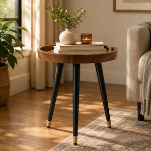May include: A round wooden side table with a raised edge and three tapered black legs featuring gold-toned metal caps. The table surface holds a white ceramic vase with green foliage, two stacked white books, and a small amber glass candle holder.