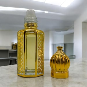 May include: A gold-coloured perfume bottle with a decorative pattern and a separate, matching cap. The bottle is filled with a light yellow liquid. The design features a geometric pattern and a clear, cylindrical rollerball top.
