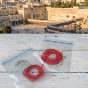 Può includere: Due sacchetti di plastica trasparente con chiusure a zip blu. Ogni sacchetto contiene uno spago rosso arrotolato e un ciondolo decorativo. Un ciondolo è una mano di Hamsa color argento, e l'altro è una mano di Hamsa color oro. Lo sfondo mostra una città con edifici in pietra.