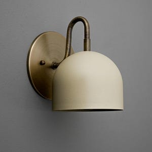May include: A wall-mounted light fitting featuring a cream-coloured, domed shade. It has a brass-toned circular backplate and a curved arm. The design is minimalist and modern, suitable for interior lighting, with a focus on functionality.