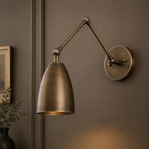 Puede incluir: Un aplique de pared con acabado en latón antiguo, pantalla cónica y brazo articulado ajustable. La lámpara incluye una placa de pared circular y una articulación giratoria en la parte superior de la pantalla para orientar la luz.