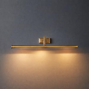 May include: A brass-toned picture light fixture. The light has a rectangular wall mount and a horizontal bar with a small adjustable arm. The light is illuminating the wall below. The fixture is a warm gold color.