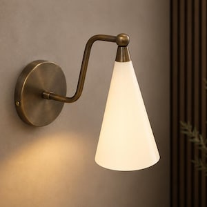 Peut inclure: Une applique murale composée d'une plaque de fixation circulaire en laiton antique, d'un bras métallique courbé et d'un abat-jour conique en verre blanc orienté vers le bas. La lampe diffuse une lueur douce sur la finition métallique.