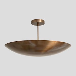 May include: A ceiling light fitting featuring a wide, shallow, bowl-shaped shade with a brushed bronze finish. The shade is suspended by a thin, vertical metal rod connected to a circular mounting canopy at the top, creating a sleek profile.