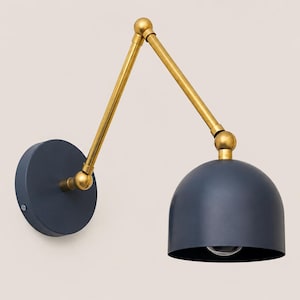 May include: A wall-mounted lamp featuring a matte dark blue circular base and a matching dome-shaped shade. The fixture includes an adjustable brass-toned metal arm with two hinged joints, connecting the base to the light shade.
