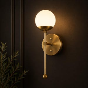 May include: A wall-mounted light fixture featuring a spherical white glass globe shade supported by a slender brass-toned metal arm. The fixture includes a circular brass wall plate and a vertical rod that extends downward, ending in a small sphere.