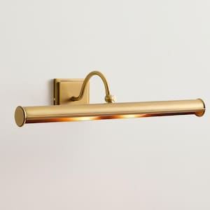 May include: A brass picture light fixture mounted on a white wall. The fixture has a rectangular base, a curved arm, and a cylindrical light housing. The light housing has a warm, golden glow.